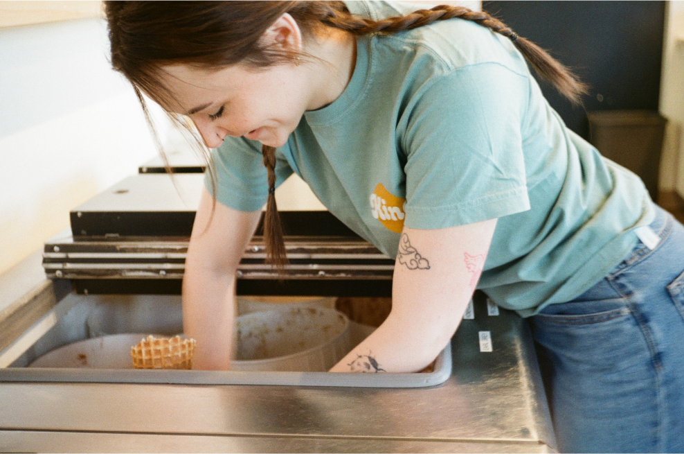 a young woman with long braieded hair in a blue shirt and jeans leans over an ice cream cooler